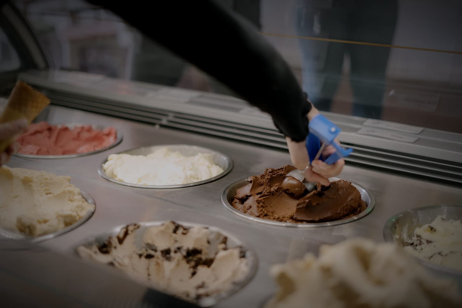 A hand in a black sleeve scoops chocolate gelato from a tub on a shop counter.